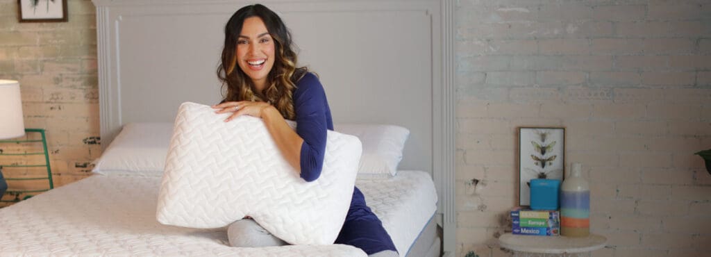 A smiling woman wearing blue with a big pillow on a bed